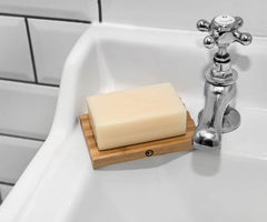 A rectangular bar of soap rests on a Bamboo Soap Dish at the edge of a white sink near a chrome faucet, with white tile and black grout behind—perfect for extending soap life and keeping grooming essentials organized.