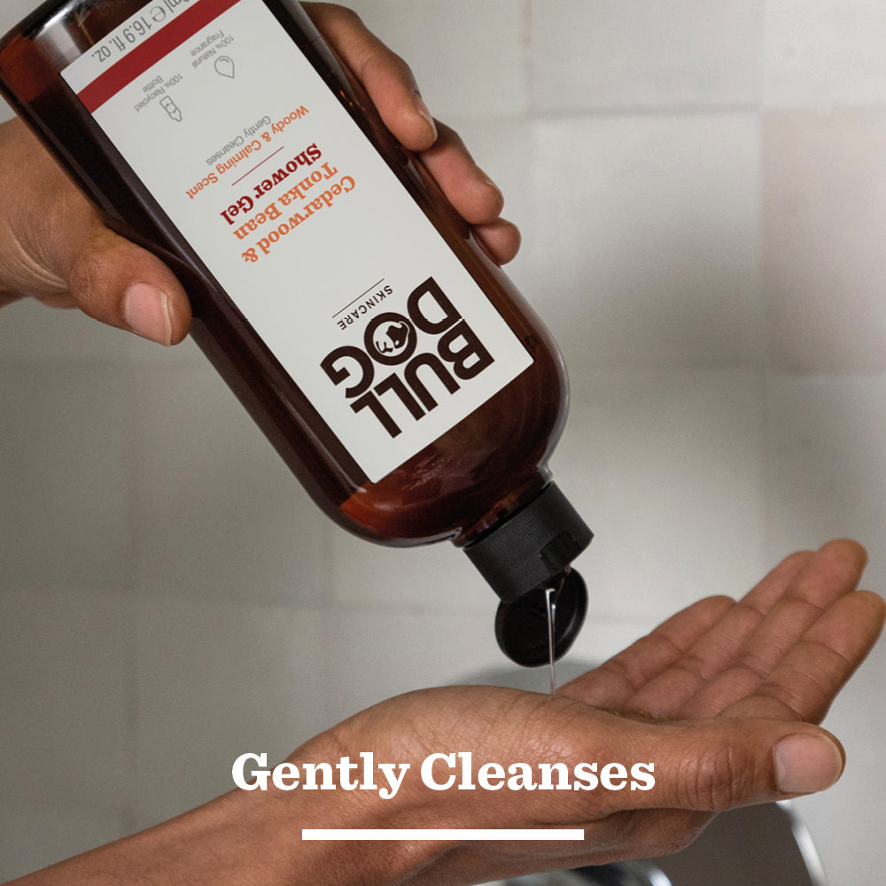 A person pours Cedarwood & Tonka Bean Shower Gel into their hand over a sink. The brown bottle with a white label reads Gently Cleanses at the bottom and emphasizes its natural fragrance.
