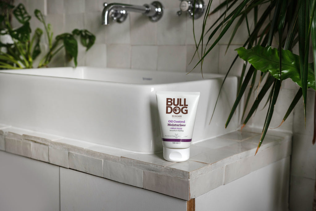 A tube of Bulldog Oil Control Face Wash with natural origin ingredients sits on the edge of a white bathroom sink, surrounded by green plants.