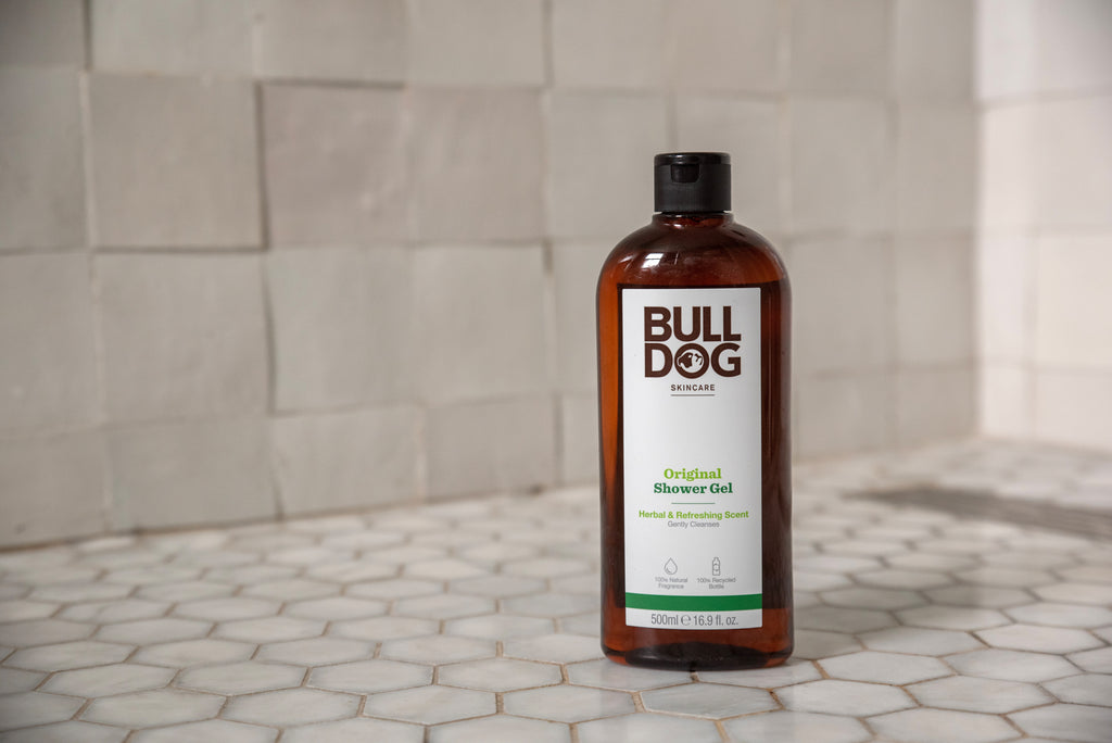 A bottle of Original Shower Gel stands on a tiled bathroom floor with gray and white hexagonal tiles and a beige tiled wall in the background, offering a refreshing scent with natural ingredients.