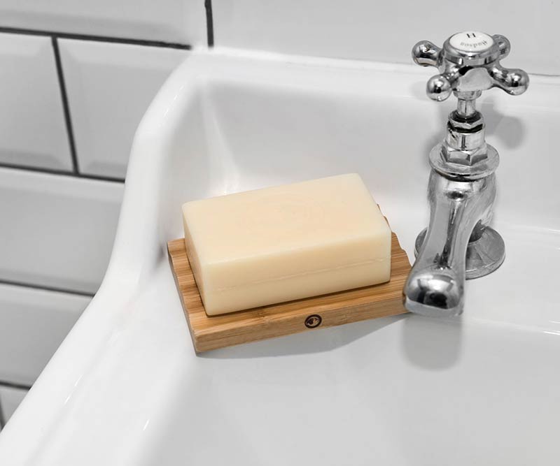 A rectangular bar of soap rests on a Bamboo Soap Dish at the edge of a white sink near a chrome faucet, with white tile and black grout behind—perfect for extending soap life and keeping grooming essentials organized.